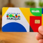 Mão segurando um cartão do programa Bolsa Família com design colorido (amarelo, azul, verde e vermelho) e chip visível, em destaque contra um fundo desfocado de cozinha com frutas e uma xícara.